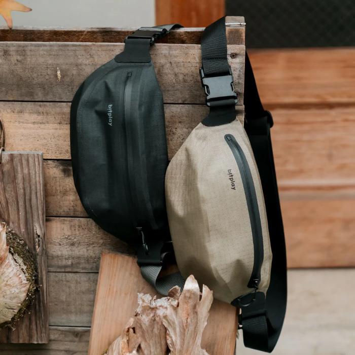 Two Bitplay AquaSeal IPX7 Waterproof Sling Bag with Strap Adapter, one black and one sand beige, on a wooden surface with natural elements.