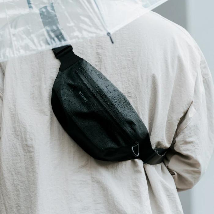 Black Bitplay AquaSeal IPX7 Waterproof Sling Bag with Strap Adapter on a beige garment with a transparent umbrella in the background